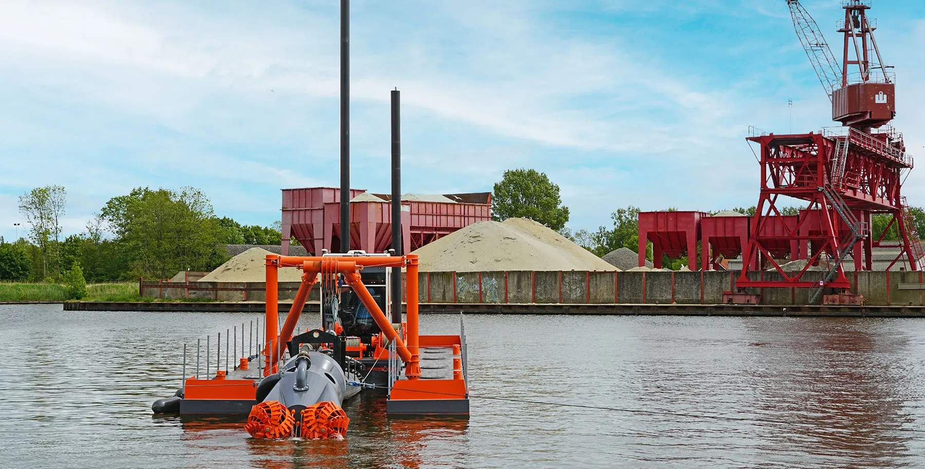 Una draga de succión con cortador dredger , con el chasis de color naranja y el cabezal de corte, flota en un canal, con una grúa pórtico roja y montones de arena y grava al fondo, en un polígono industrial.
