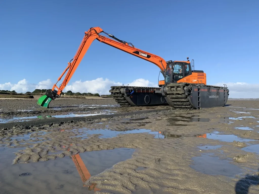 Excavadora anfibia Bell con brazo de gran alcance y cuchara verde trabajando en las marismas durante la marea baja, equipada con orugas tipo « pontoon » para acceder a los humedales.