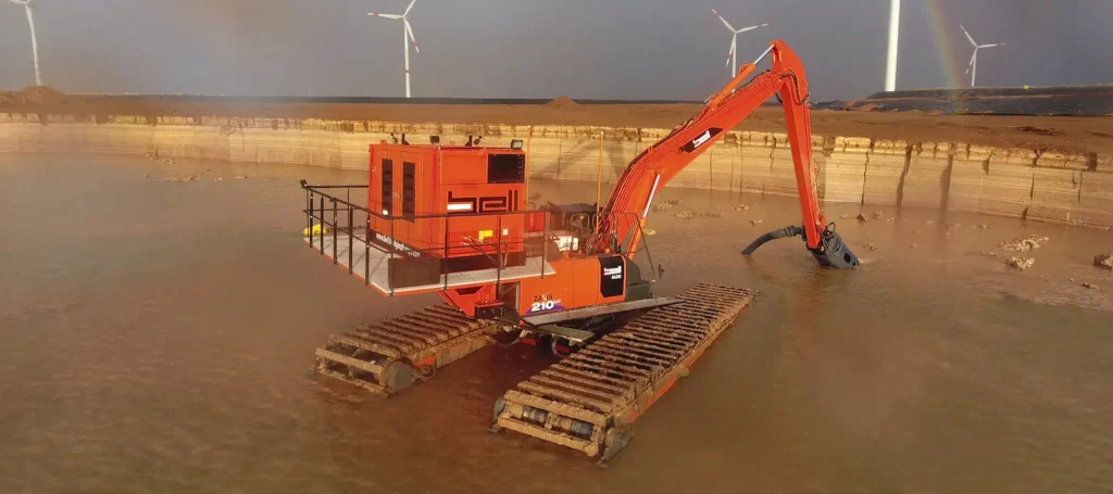 La excavadora anfibia « dredging » de Bell Dredging Pumps se encuentra en un embalse, equipada con un cabezal de corte sumergido, con aerogeneradores al fondo.