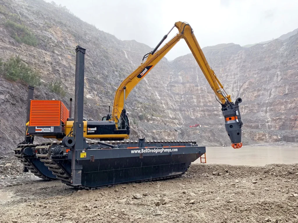 Excavadora anfibia Bell BAE300 dredging con brazo articulado alargado y accesorio cortador, situada en un terraplén arenoso cerca de un estanque de cantera, con paredes rocosas de excavación al fondo.