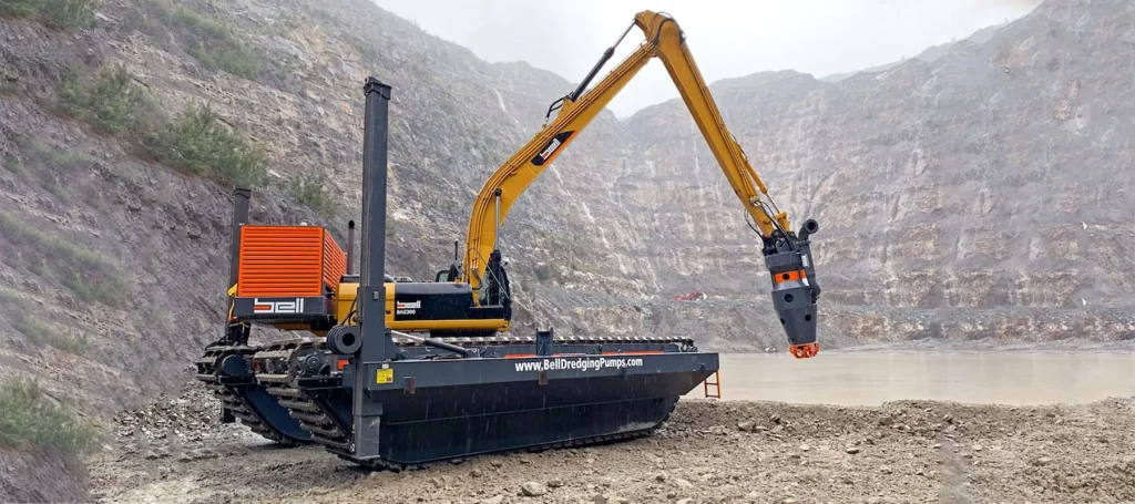 Máquina de tipo excavadora anfibia « dredging », con base sobre orugas pontoon y una pluma amarilla extendida equipada con un cabezal de corte, que opera en una mina a cielo abierto o cantera rodeada de muros rocosos en terrazas.
