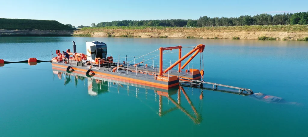 dredger e flotante de succión con cortador, con la estructura de la grúa y la cabina de mando de color naranja, anclado en aguas tranquilas de color turquesa, con una costa arenosa y una línea de árboles del bosque al fondo.