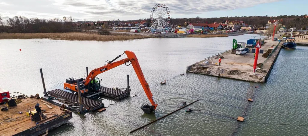 Vista aérea de una excavadora hidráulica montada sobre una plataforma flotante pontoon realizando operaci dredging es en el canal de un puerto, con una noria y una localidad costera visibles al fondo.