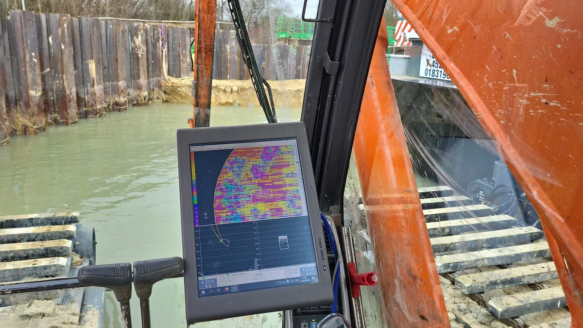 Excavator cab view with a color depth-scanning monitor displaying underwater terrain data, overlooking a water-filled dredging site enclosed by sheet piling walls.