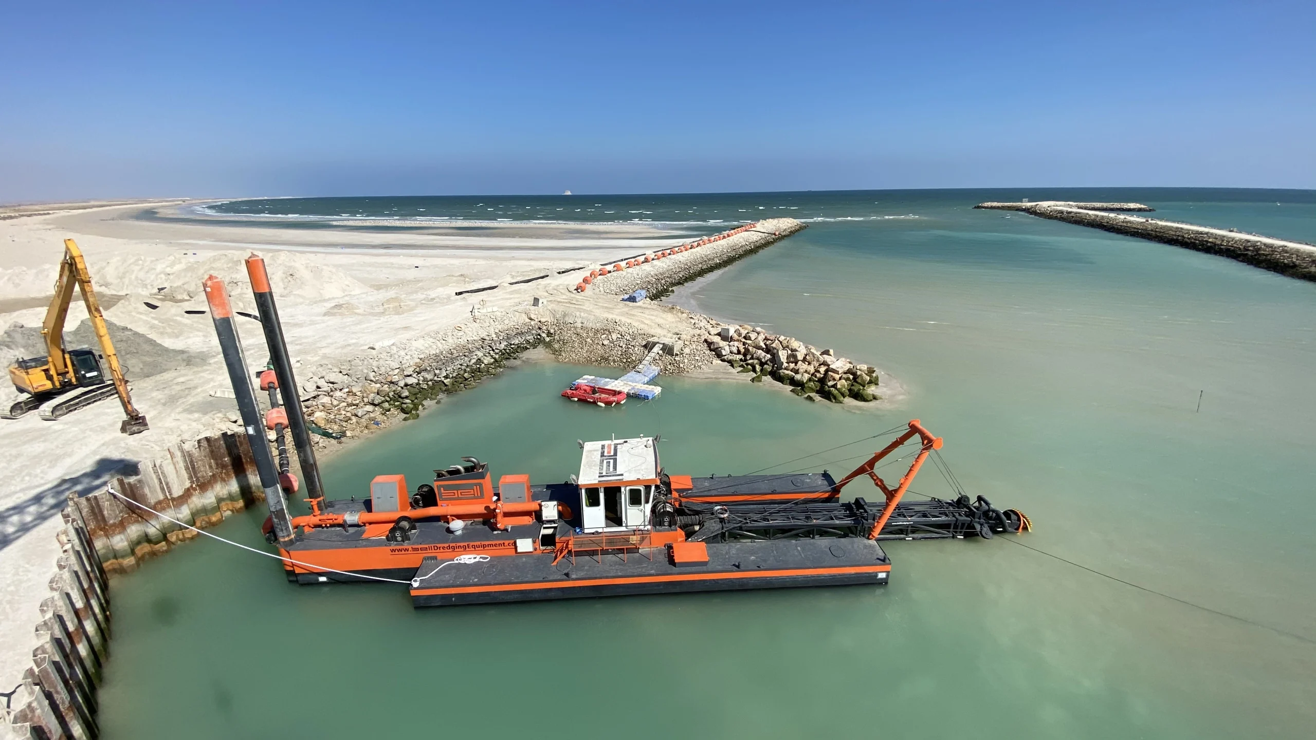 Vista aérea de una draga de succión con cuchilla de color naranja y negro dredger amarrada en el canal de un puerto, con un embarcadero, un rompeolas, una excavadora y un litoral arenoso visibles al fondo, bajo un cielo azul y despejado.